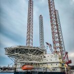A towering offshore oil rig platform situated dockside against the blue ocean and cloudy sky.